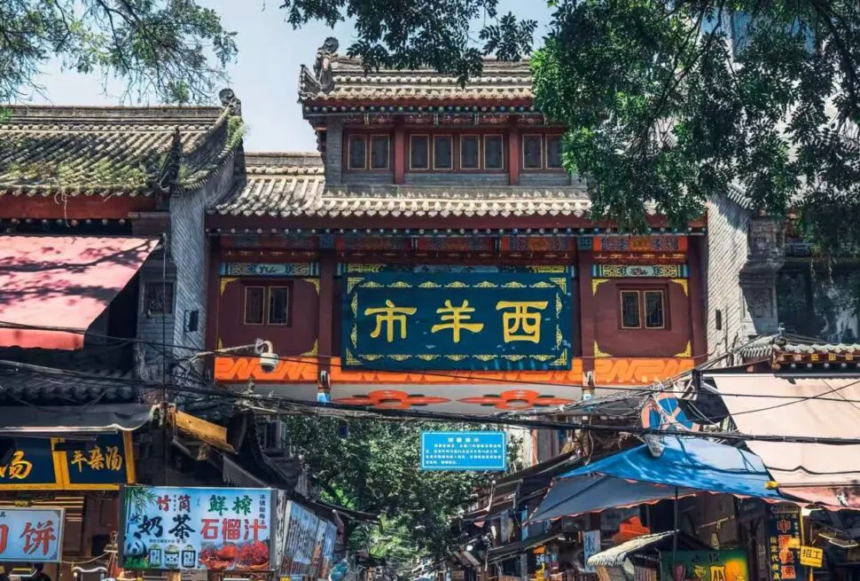 Decorative Chinese gateway over a bustling market street with shops and stalls beneath a blue sign panel and ornate roof.