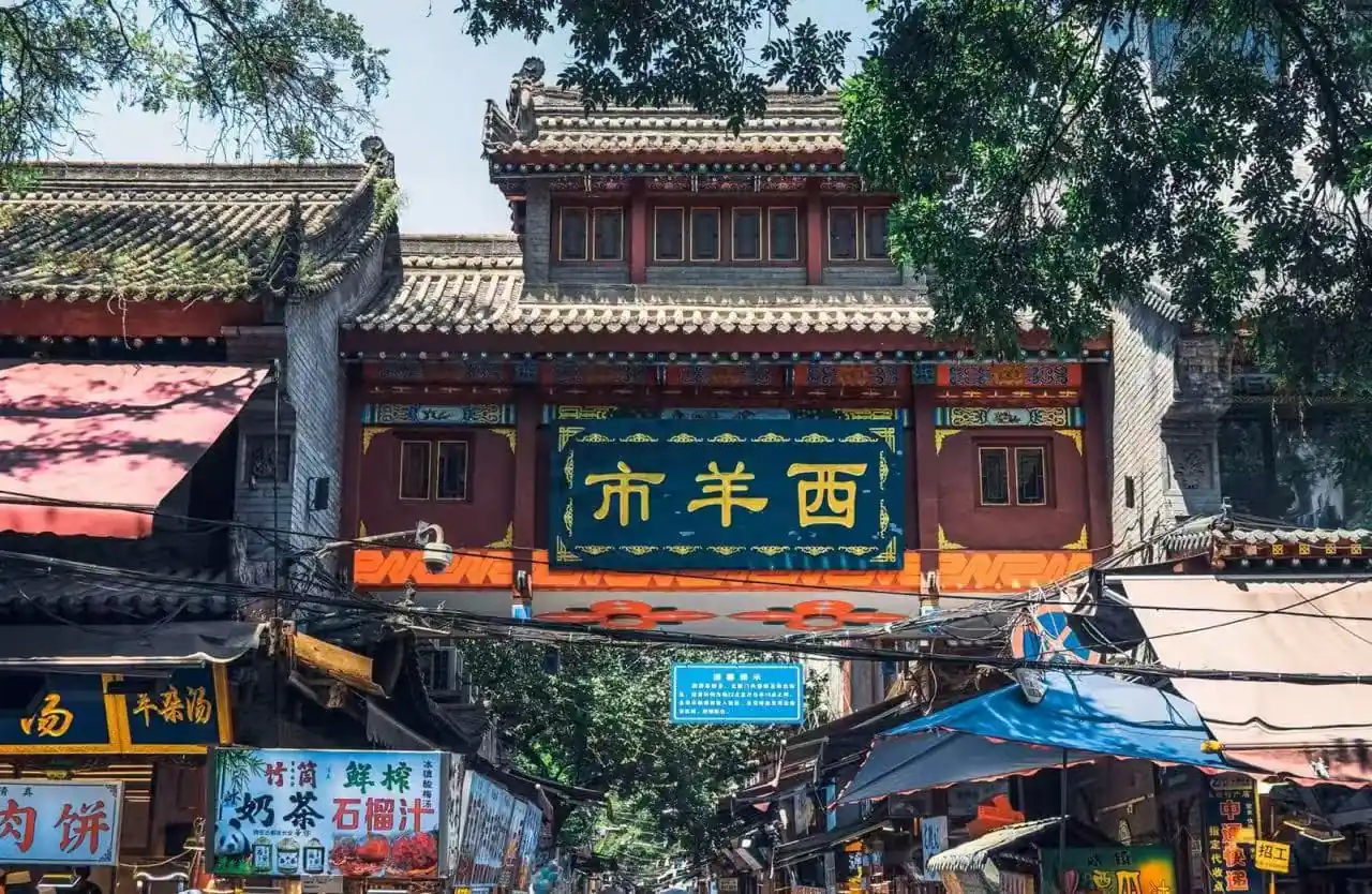 Decorative Chinese gateway over a bustling market street with shops and stalls beneath a blue sign panel and ornate roof.