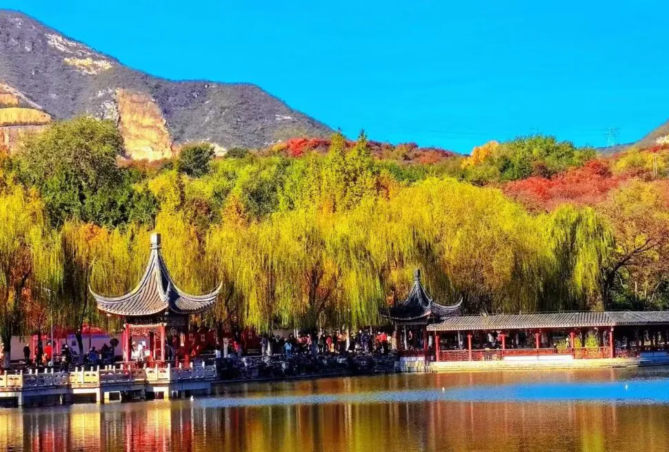Lakeside scene with a traditional Chinese pavilion, yellow willows, and shoppers near a red railing against a mountainside.