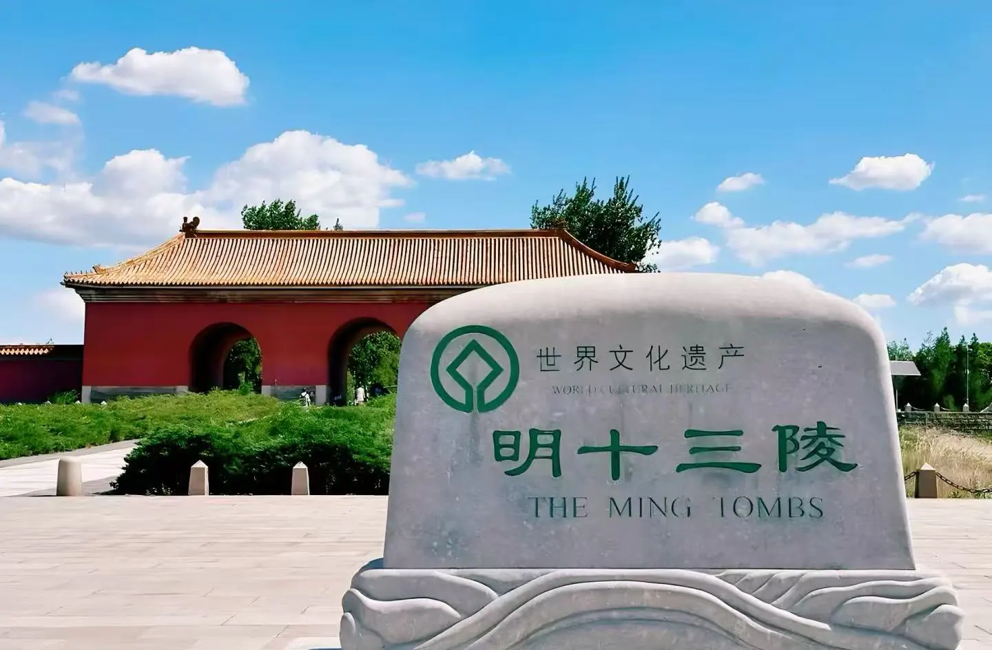 Stone monument with green emblem and Chinese text reading 'World Cultural Heritage' and 'The Ming Tombs' in front of a red gate with tiled roof under a blue sky.