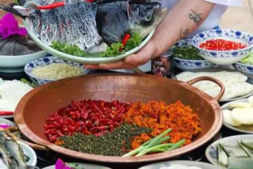 A person holds a plate of whole fish heads above a table of ingredients; in a copper bowl in the foreground are chopped red chilies, orange spices, green herbs, and scallions surrounded by bowls of condiments.