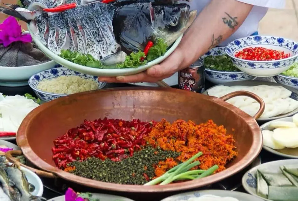 A person holds a plate of whole fish heads above a table of ingredients; in a copper bowl in the foreground are chopped red chilies, orange spices, green herbs, and scallions surrounded by bowls of condiments.