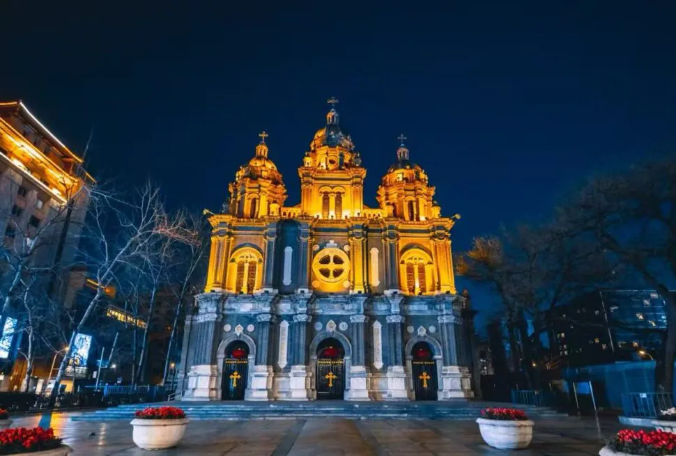 Night view of a grand historic cathedral lit in warm golden lights, featuring three domed towers and ornate façade.