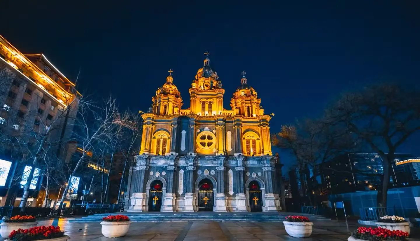 Night view of a grand historic cathedral lit in warm golden lights, featuring three domed towers and ornate façade.
