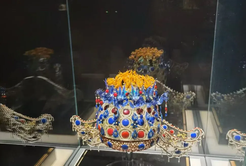 Ornate jeweled crown with blue and red gems and a yellow floral top, displayed in a glass case