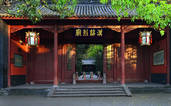 Red wooden Chinese temple gate with hanging lanterns and a courtyard beyond, under green trees