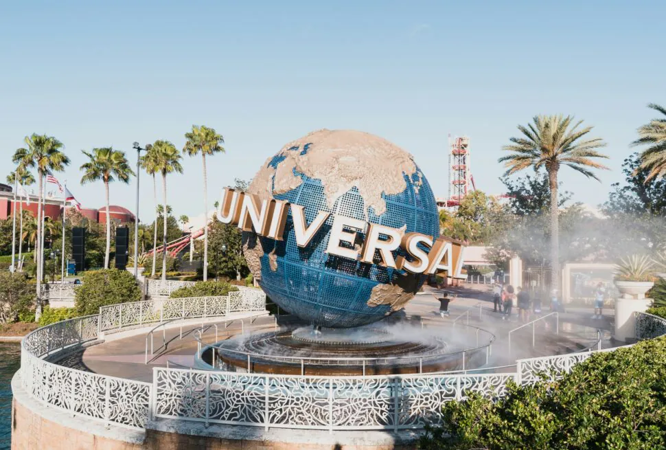 Globe sculpture with blue oceans and gold landmasses, spelling 'UNIVERSAL' in large letters, at Universal Studios theme park entrance with palm trees and mist fountain in foreground.