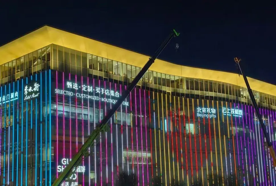 Silk Street Market,Beijing,Chinatour Night view of a glass-front shopping mall with multicolored vertical neon lights and construction cranes in front.