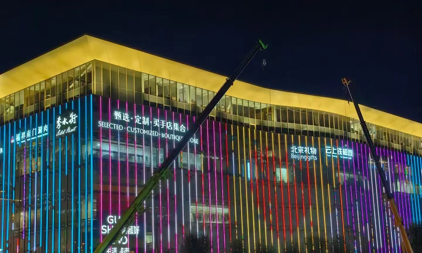 Silk Street Market,Beijing,Chinatour Night view of a glass-front shopping mall with multicolored vertical neon lights and construction cranes in front.