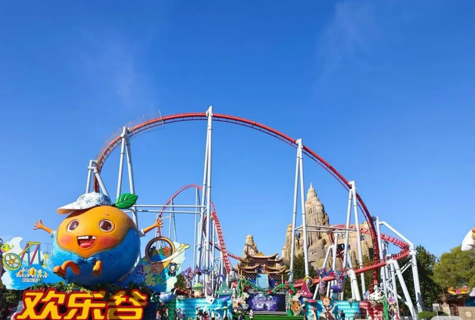 Large orange cartoon mascot with outstretched arms at a colorful amusement park, red roller coaster looping in the background under a blue sky.