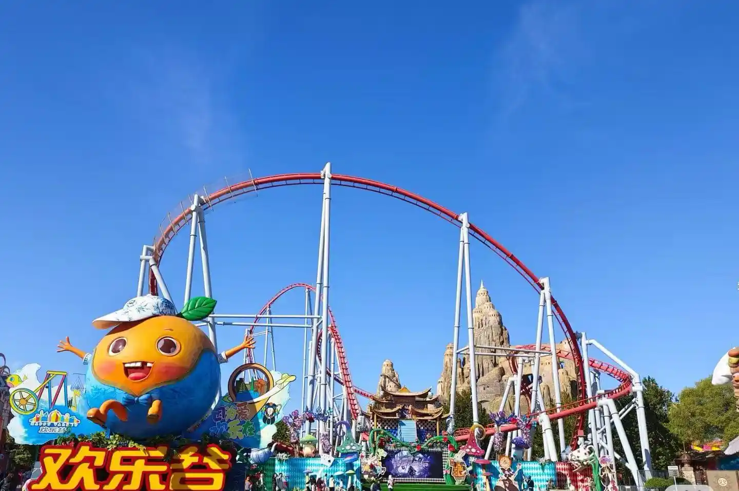 Large orange cartoon mascot with outstretched arms at a colorful amusement park, red roller coaster looping in the background under a blue sky.