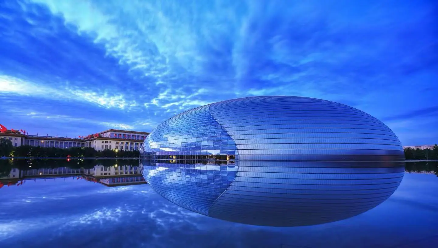 National Centre for the Performing Arts (the Egg) reflected in a calm pool under a blue sky with surrounding buildings and trees.