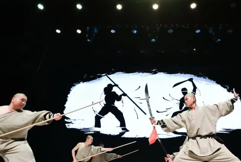 Three martial artists in beige robes wield wooden staffs and swords on a stage, dramatic action against a white brushstroke backdrop.