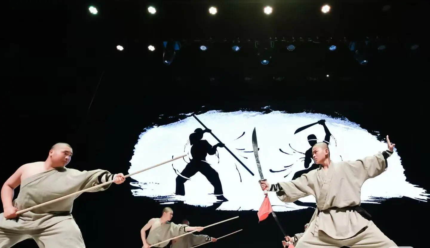 Beijing Kung Fu Show,Chinatour Three martial artists in beige robes wield wooden staffs and swords on a stage, dramatic action against a white brushstroke backdrop.