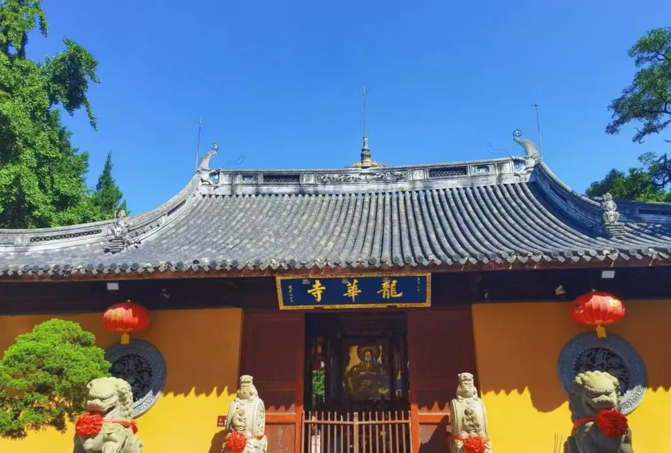 Entrance of a traditional Chinese temple with yellow walls, grey tiled roof, red lanterns, and stone guardian lions against a blue sky