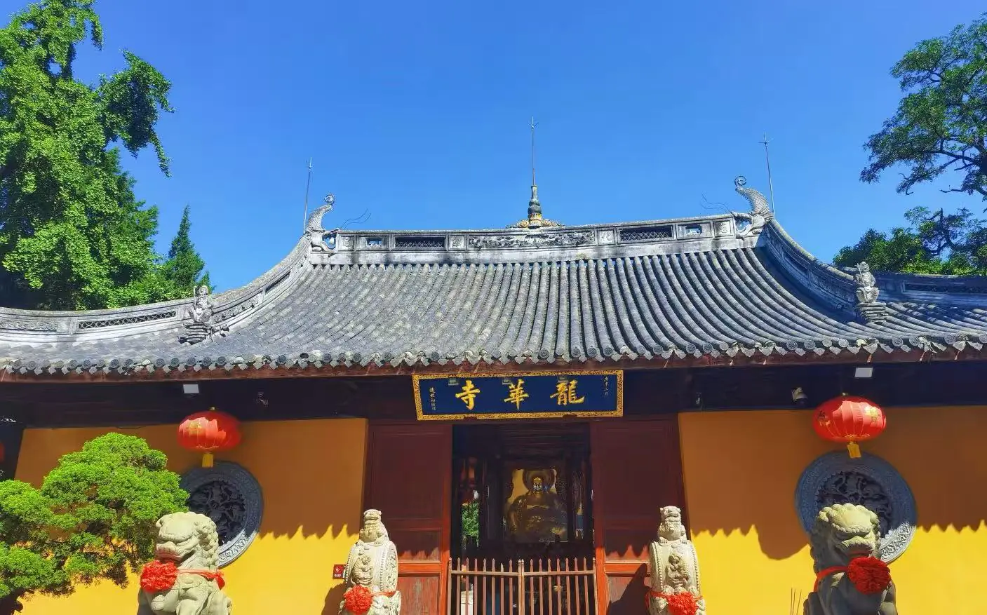 Entrance of a traditional Chinese temple with yellow walls, grey tiled roof, red lanterns, and stone guardian lions against a blue sky