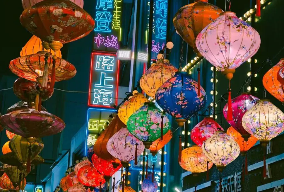 Colorful Chinese lanterns hanging above a night market street with neon signs in blue and red.