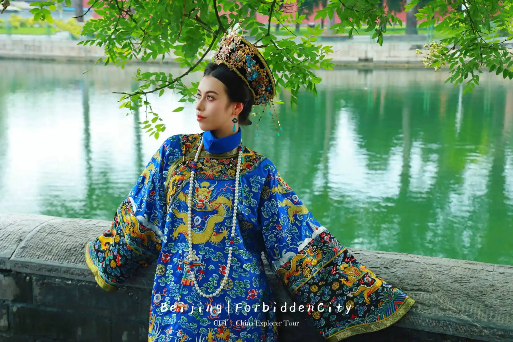 hanfu photoshoot, the forbidden city, beijing, qing dynasty, female, solo, chinatravel, Woman in an ornate blue dragon-embroidered traditional dress and elaborate headdress poses by a green lake, leaning on a stone railing (Beijing Forbidden City promotional image).