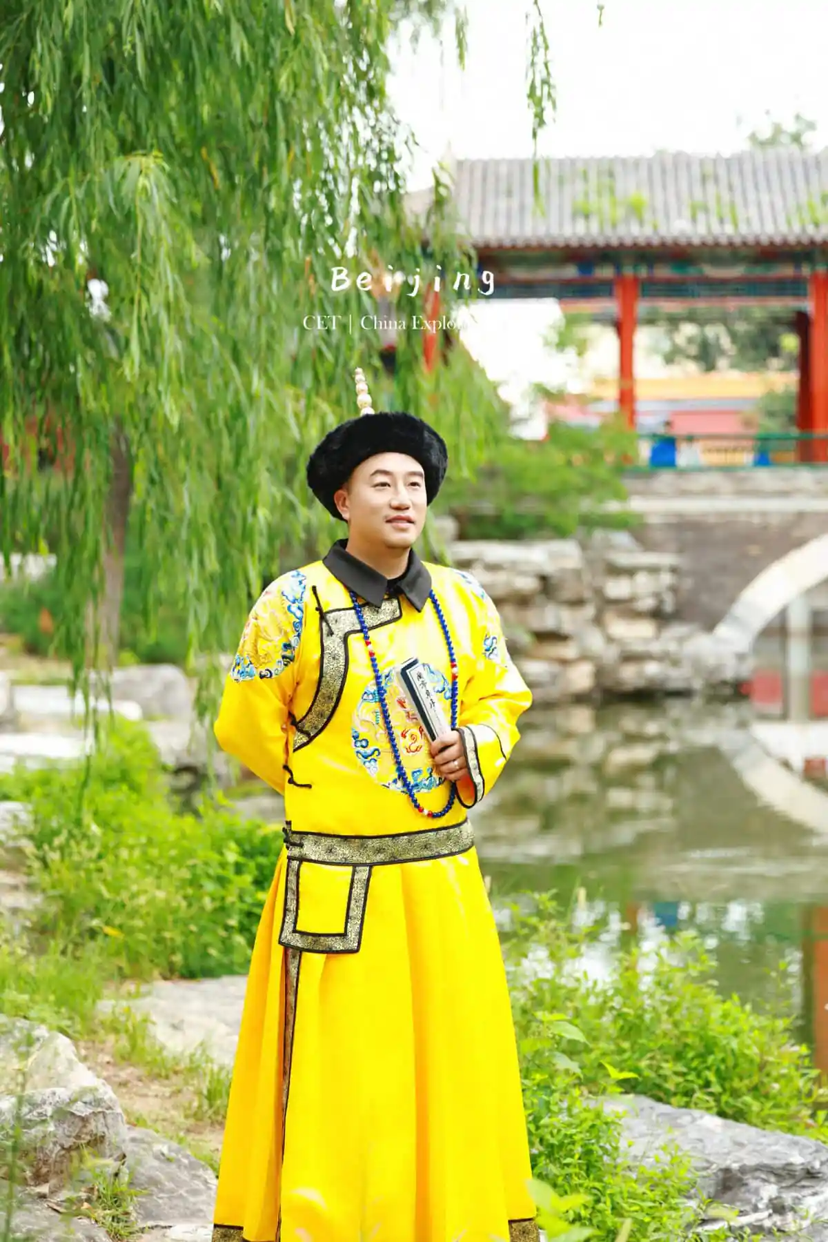 hanfu photoshoot, the forbidden city, beijing, qing dynasty, male, solo, chinatravel, Man in a bright yellow traditional Chinese robe and black hat, holding a book, standing in a garden with a willow tree and a temple-like background.