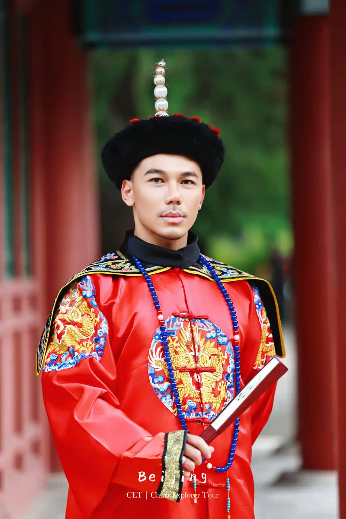 hanfu photoshoot, the forbidden city, beijing, qing dynasty, male, solo, chinatravel, Portrait of a man in traditional Chinese imperial ceremonial dress: red dragon-embroidered robe, black fur hat with pearl-topped ornament, holding a scroll outdoors.