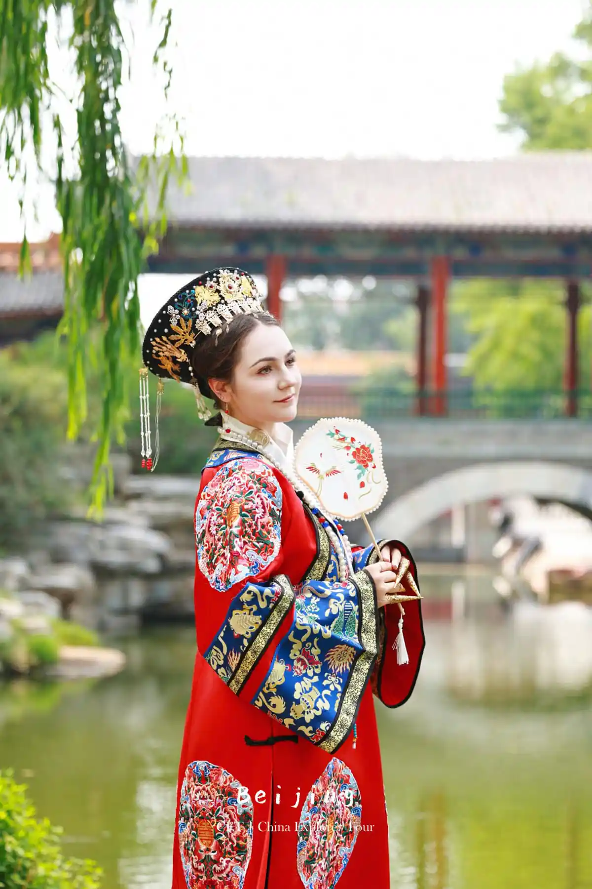 Hanfu photoshoot, zhongshan park, beijing, chinatravel, Woman in an ornate traditional Chinese headdress and red embroidered dress holding a decorative fan by a pond and bridge.
