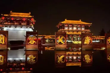 Tang Paradise Theme Zone,Xi`an,Chinatour Night scene of historic Chinese palaces lit in warm light, with red lanterns and a row of circular golden sign frames reflecting in the water.