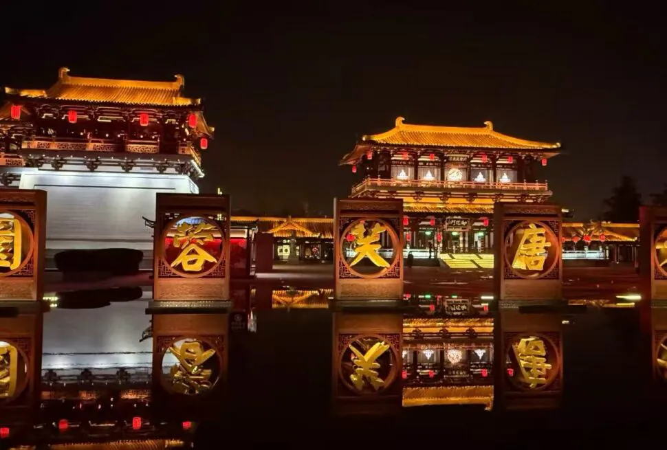 Tang Paradise Theme Zone,Xi`an,Chinatour Night scene of historic Chinese palaces lit in warm light, with red lanterns and a row of circular golden sign frames reflecting in the water.