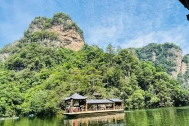 Wooden pavilion-style boat on a calm green river, with forested cliffs and a blue sky above.