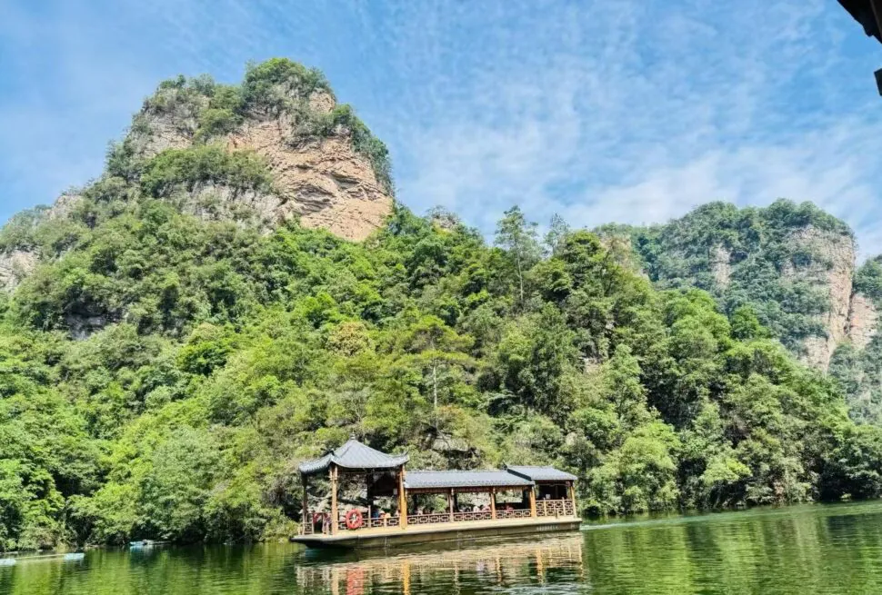 Baofeng Lake,Zhangjiajie,Chinatour Wooden pavilion-style boat on a calm green river, with forested cliffs and a blue sky above.