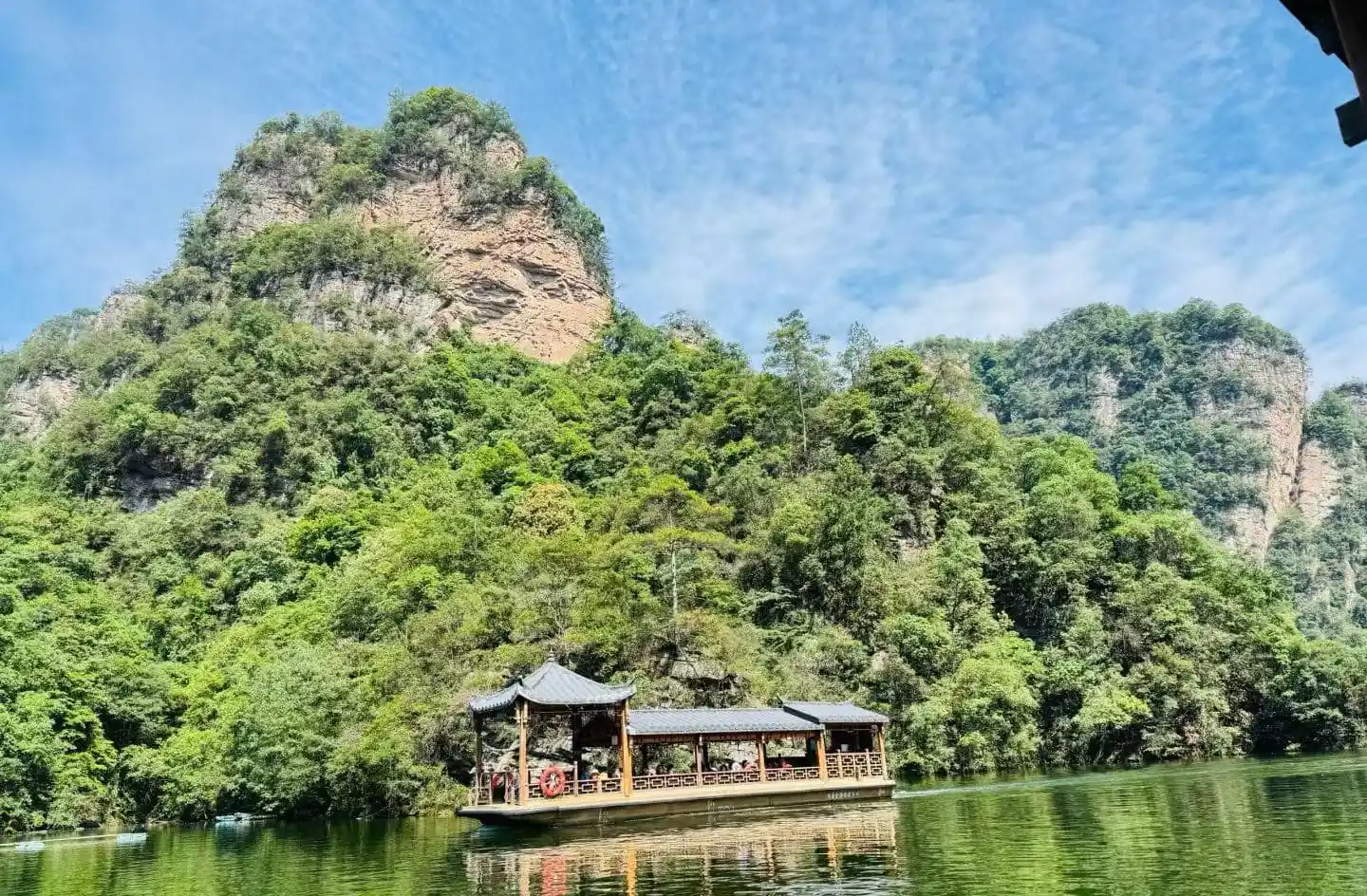 Baofeng Lake,Zhangjiajie,Chinatour Wooden pavilion-style boat on a calm green river, with forested cliffs and a blue sky above.