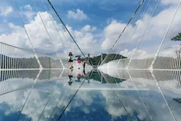 People walk on a glass-bottom bridge with a clear blue sky, their reflections visible in the mirrored surface below.