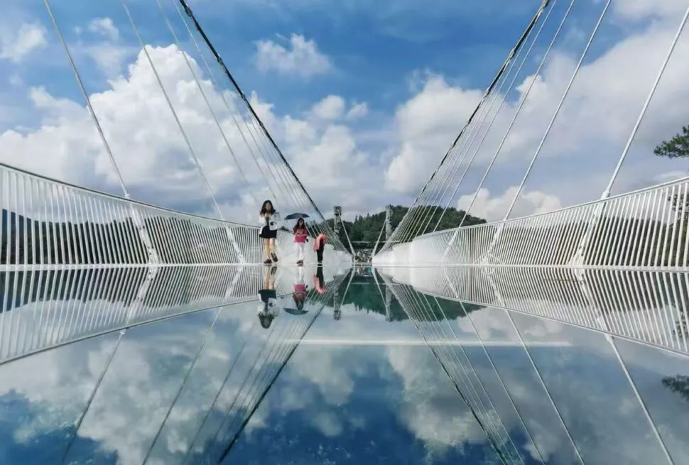 People walk on a glass-bottom bridge with a clear blue sky, their reflections visible in the mirrored surface below.