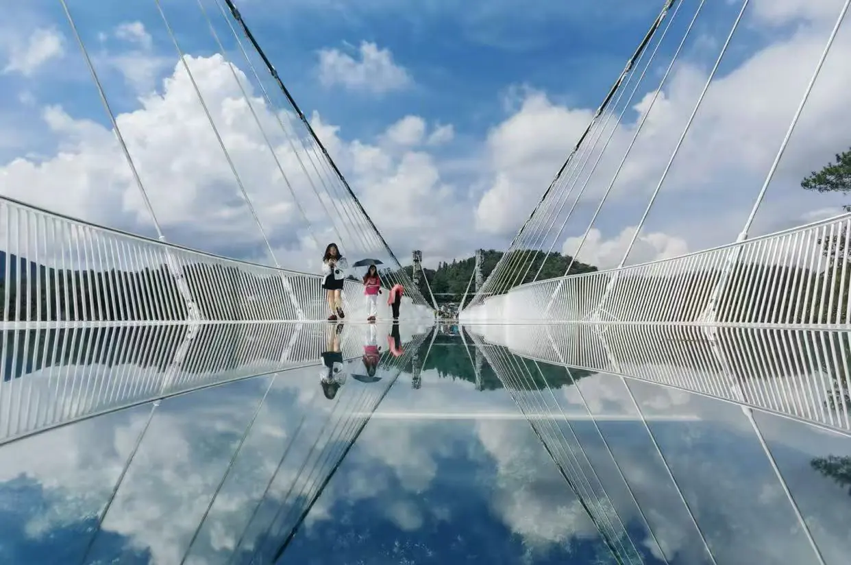 Tianmen Mountain Cableway and Glass Skywalk,Zhangjiajie,Chinatour People walk on a glass-bottom bridge with a clear blue sky, their reflections visible in the mirrored surface below.