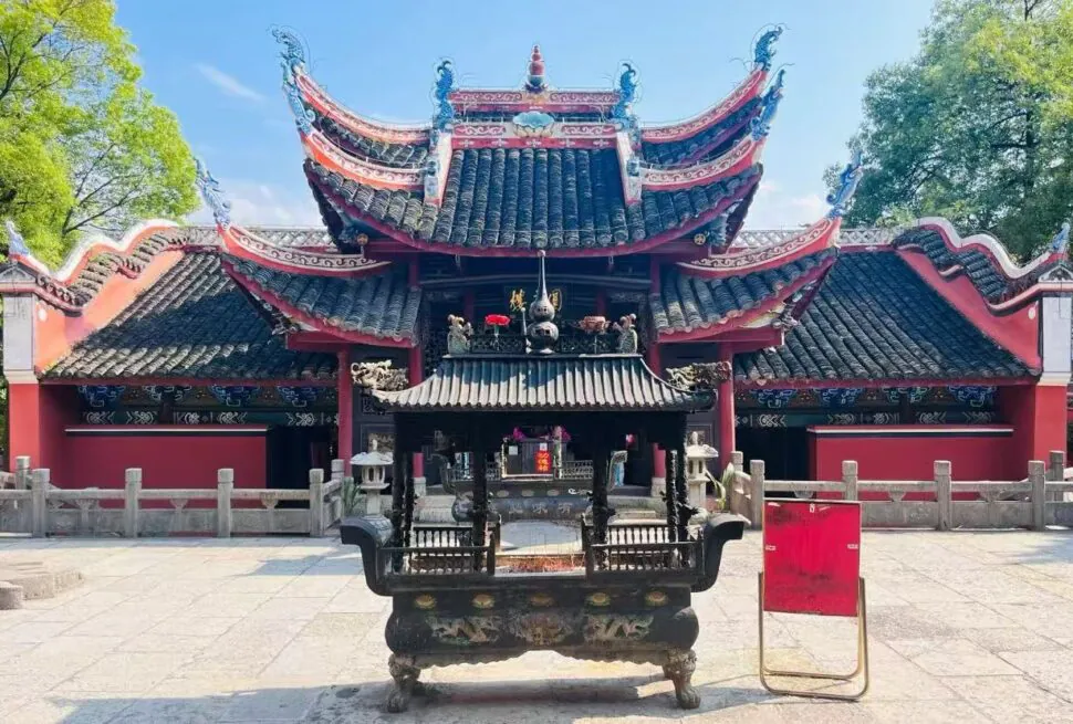 Old-style Chinese temple with ornate curved roofs and red walls, seen from a large ceremonial incense burner in the foreground.