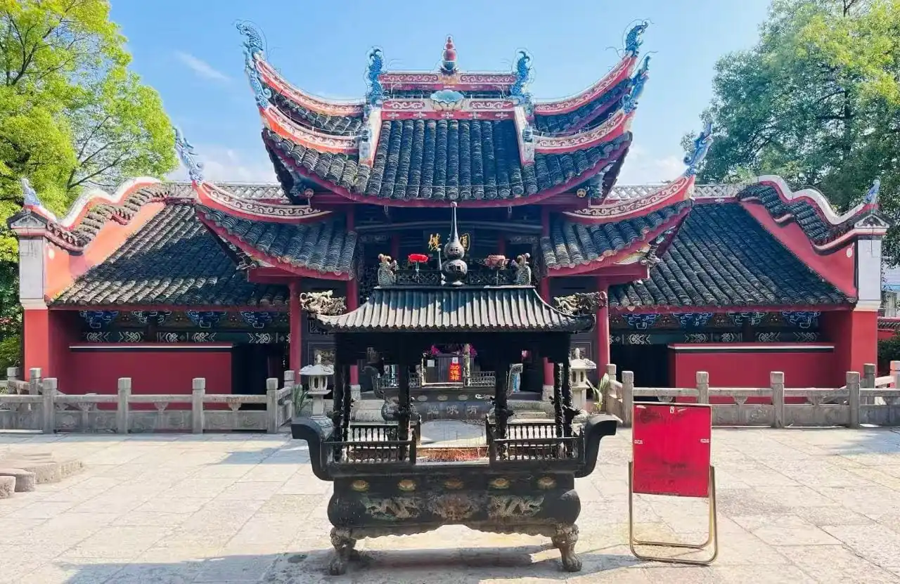 Puguang Temple,Zhangjiajie,Chinatour Old-style Chinese temple with ornate curved roofs and red walls, seen from a large ceremonial incense burner in the foreground.