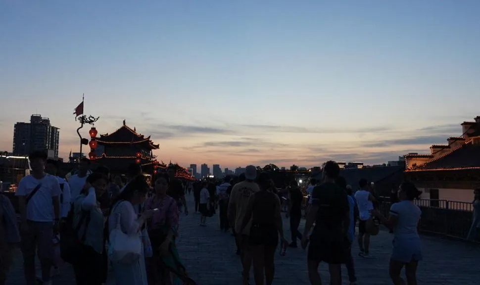Crowd strolling along a waterfront at dusk, with a pagoda-style building and red lanterns lit along the walkway.