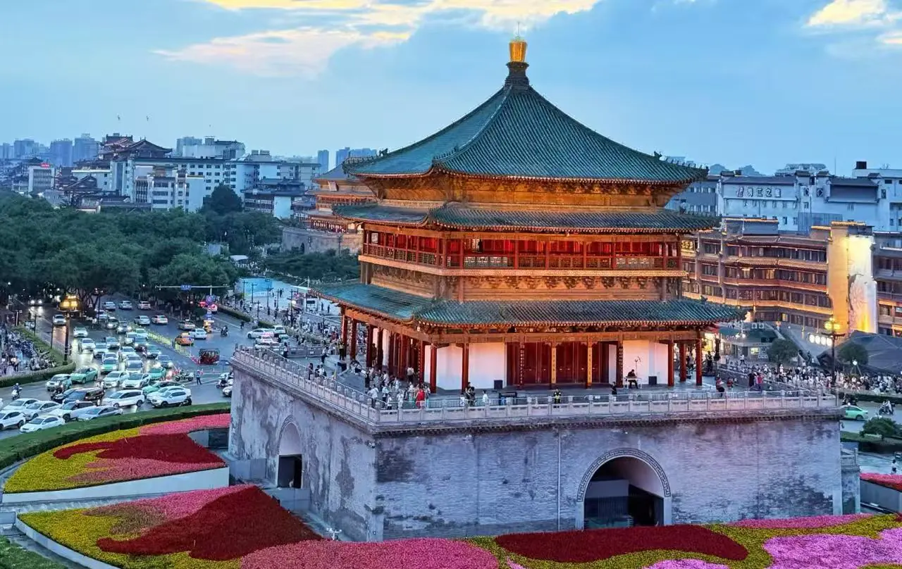 Traditional multi-tier Chinese pagoda atop a stone fortress overlooking a city, with colorful flowerbeds in the foreground.