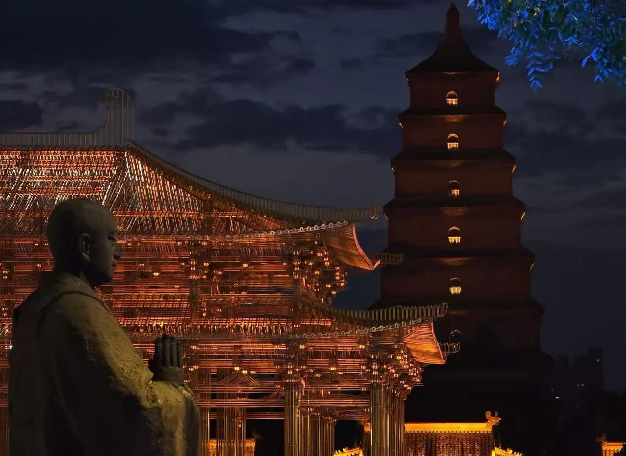 Night scene of a lit Chinese temple pavilion with a tall pagoda and a stone statue in the foreground at night.