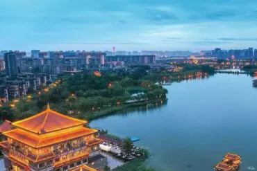 Qujiang Pool Park,Xi`an,Chinatour Aerial view of a city by a large lake, with a glowing, multi-tiered pagoda on the bank and skyline in the distance at dusk.