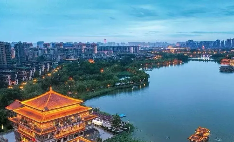 Qujiang Pool Park,Xi`an,Chinatour Aerial view of a city by a large lake, with a glowing, multi-tiered pagoda on the bank and skyline in the distance at dusk.