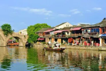 Stone arch bridge over a calm canal, with green trees and traditional riverside shops lining the water’s edge; a decorated boat glides by.