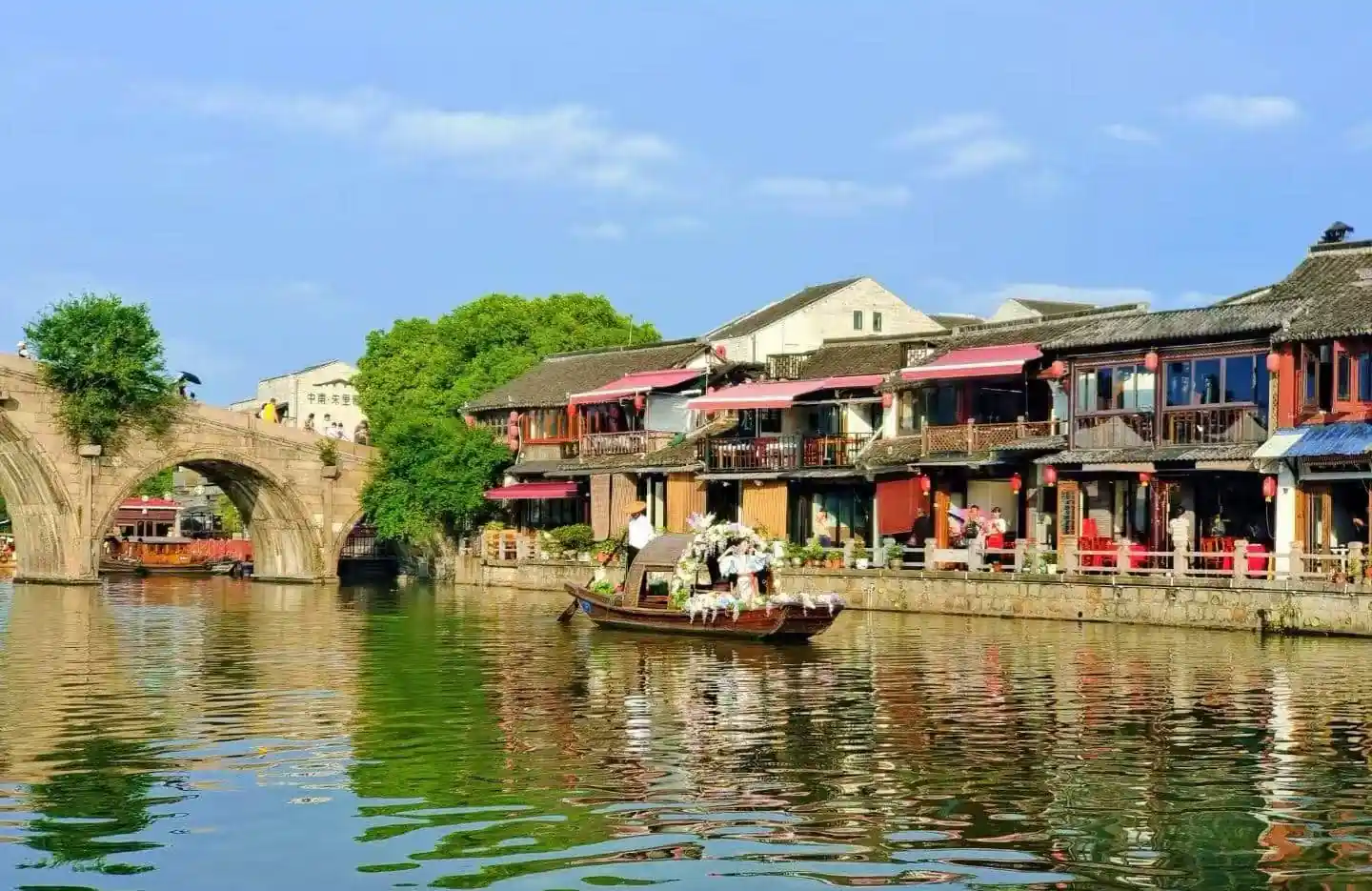 Zhujiajiao Water Town,Shanghai,Chinatour Stone arch bridge over a calm canal, with green trees and traditional riverside shops lining the water’s edge; a decorated boat glides by.