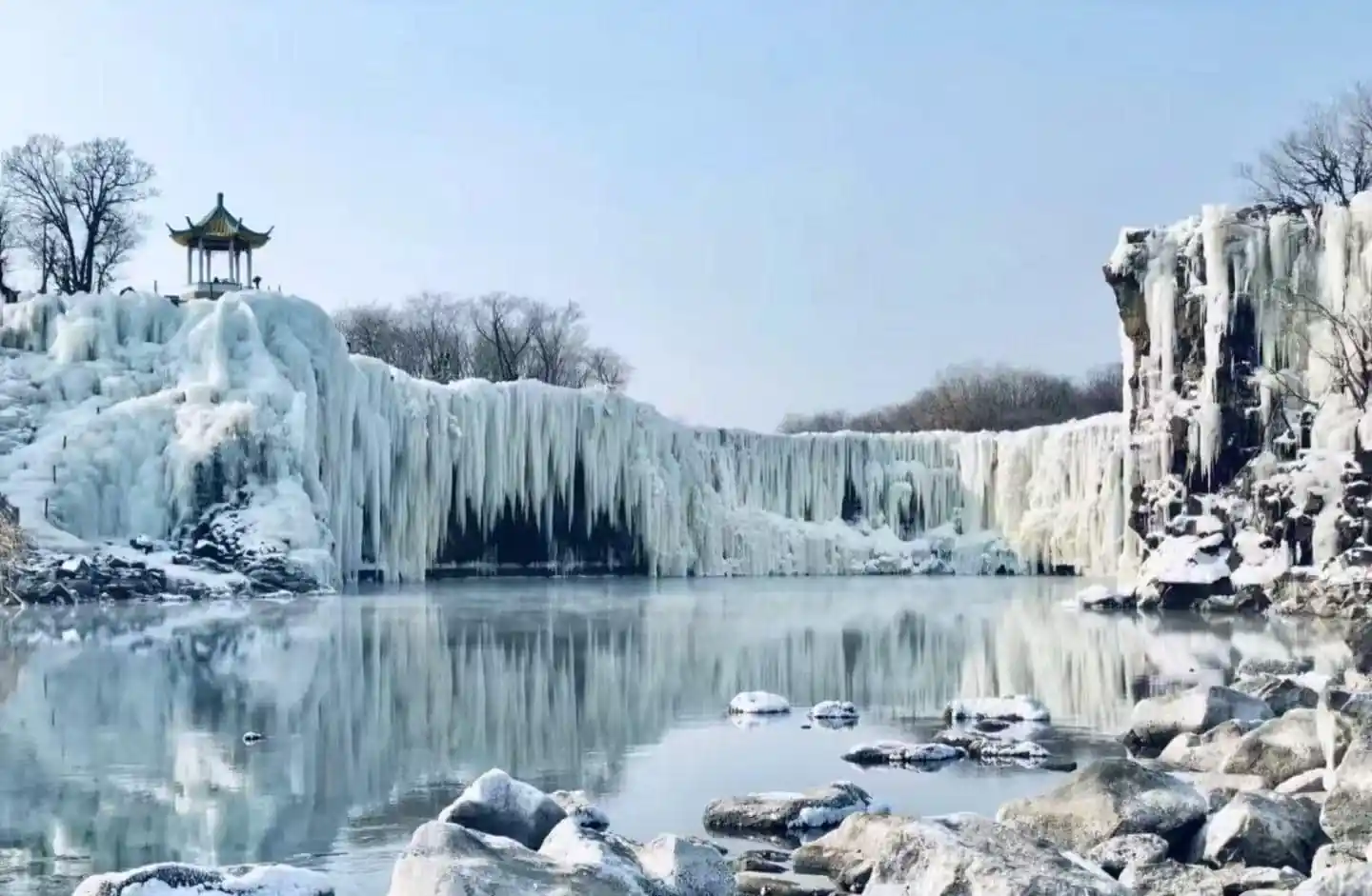 Jingpo Lake and Diaoshuilou Waterfall,Mudanjiang,Chinatour Winter scene with a frozen waterfall and icicles, a small pagoda on the left, and a calm, reflective lake below.