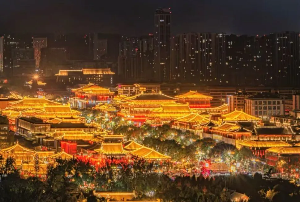 Datang Everbright City,Xi`an,Chinatour Nighttime panorama of a brightly lit traditional Chinese palace complex with glowing orange rooftops against a modern city skyline.