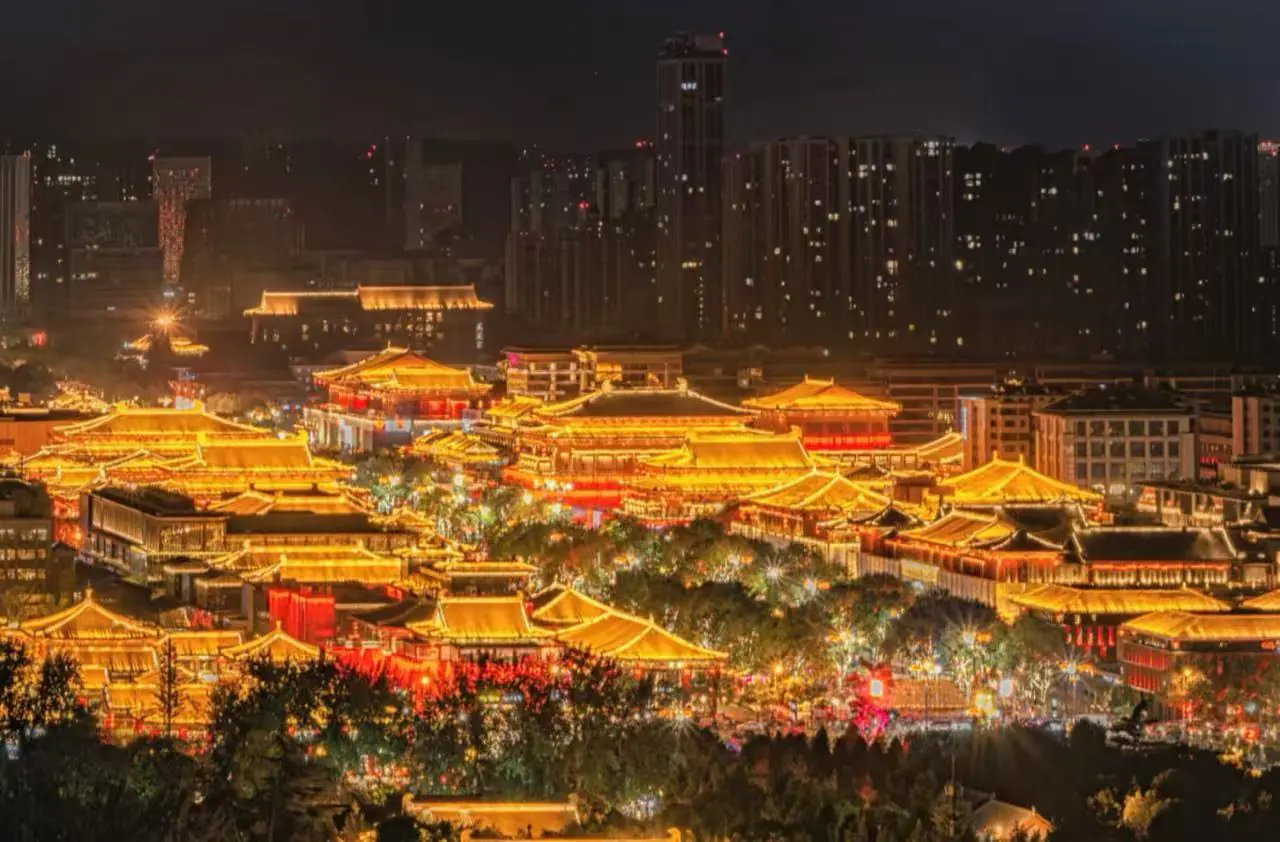 Nighttime panorama of a brightly lit traditional Chinese palace complex with glowing orange rooftops against a modern city skyline.