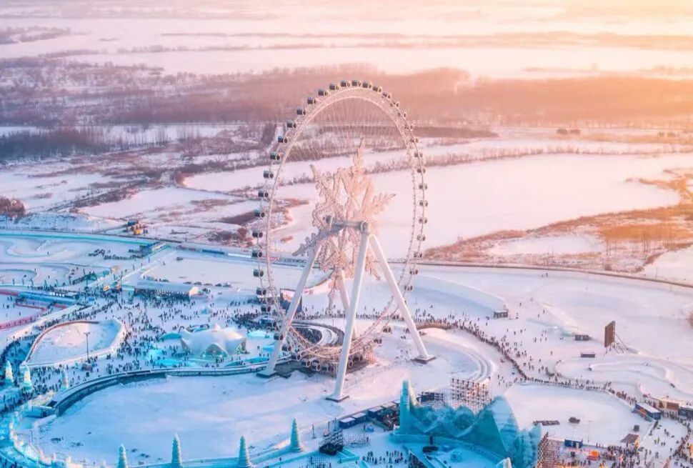 Aerial view of a large, illuminated Ferris wheel in a snow-covered amusement park at sunset, with crowds below.