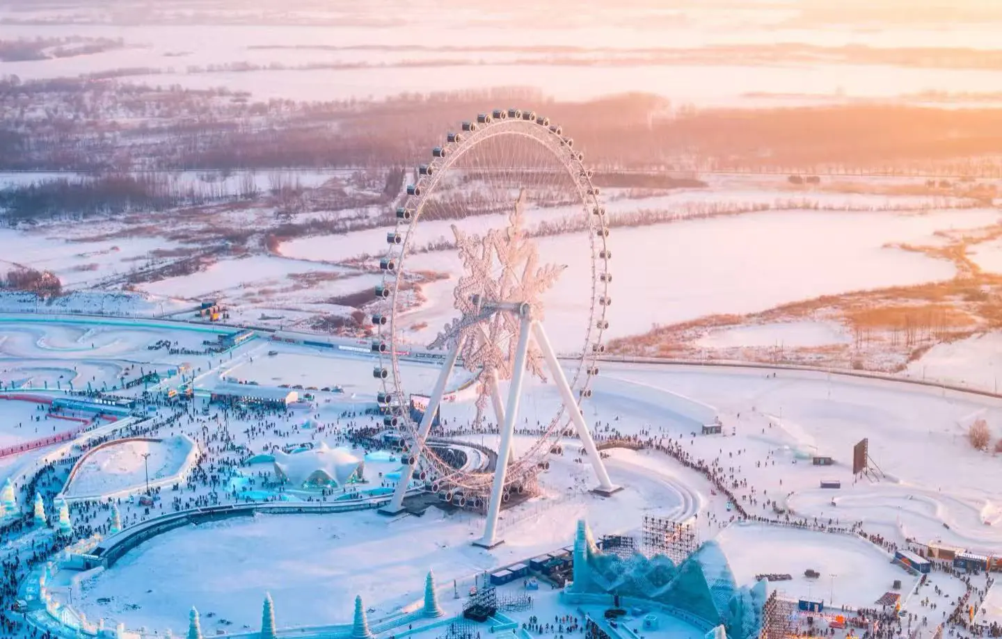 Aerial view of a large, illuminated Ferris wheel in a snow-covered amusement park at sunset, with crowds below.