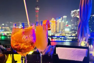Two cocktails on a rooftop bar at night with a glowing city skyline in the background.