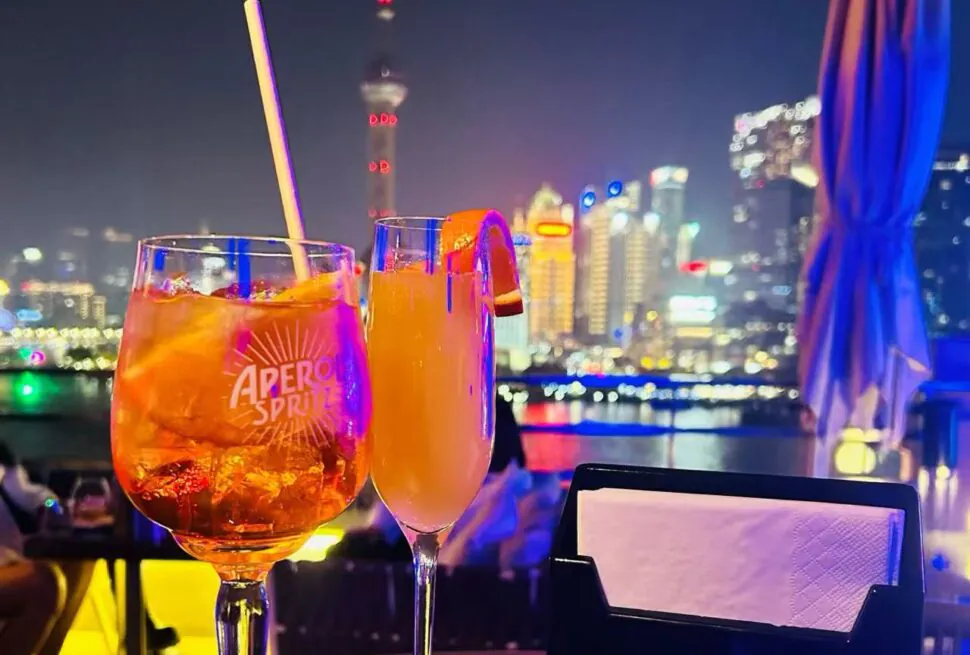 Two cocktails on a rooftop bar at night with a glowing city skyline in the background.