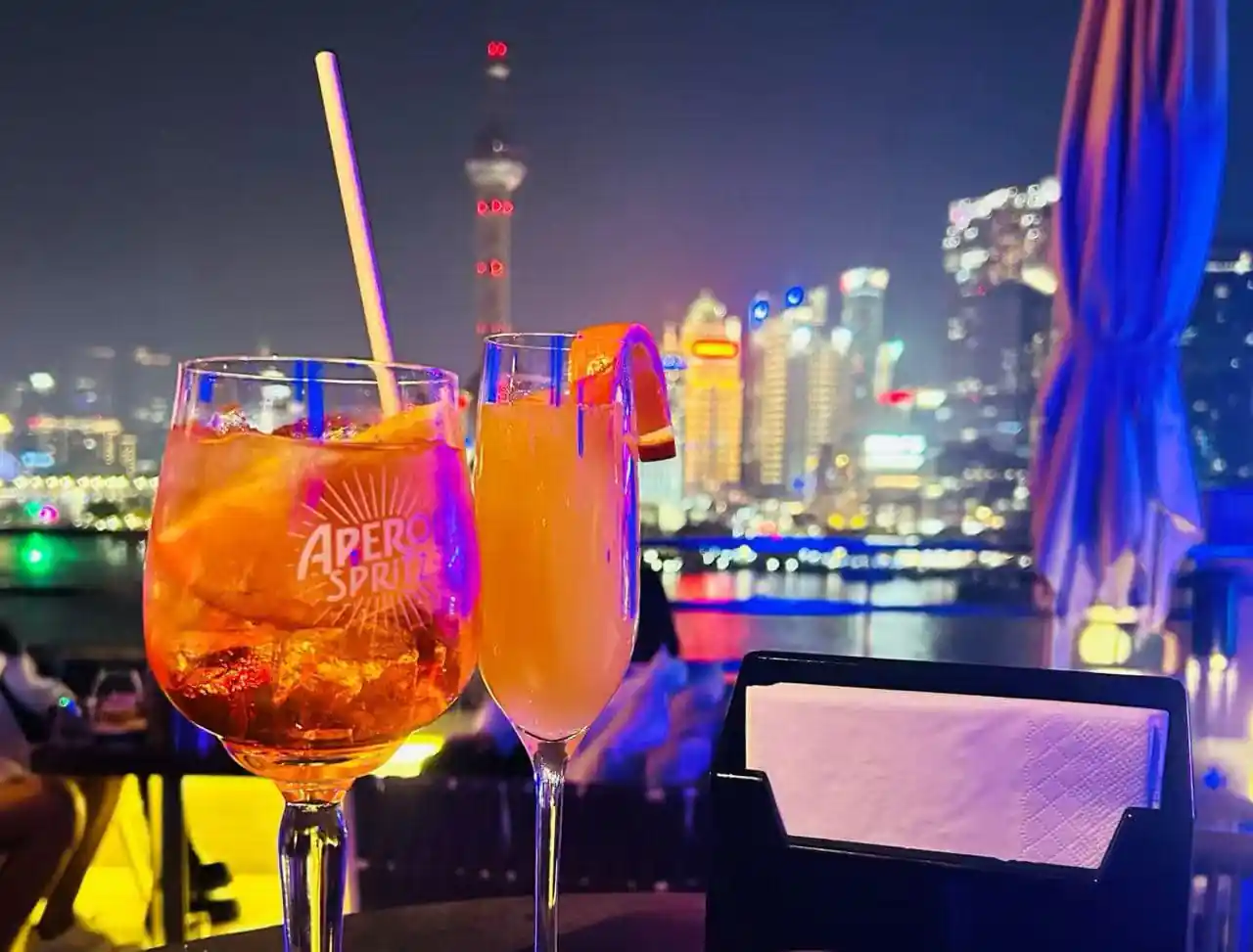 Two cocktails on a rooftop bar at night with a glowing city skyline in the background.